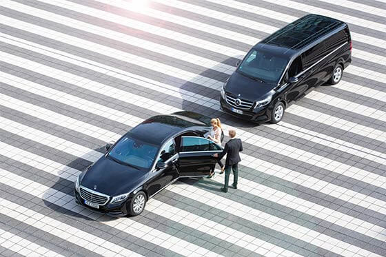 Ein Chauffeur öffnet die Tür einer Limousine und hilft einer Frau beim einsteigen. Dahinter wartet ein Minivan.